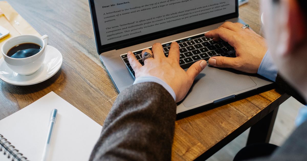 Close-up over-the-shoulder view of a person in a blazer typing on a laptop at a wooden desk, with a cup of black coffee, a notepad and pen beside them