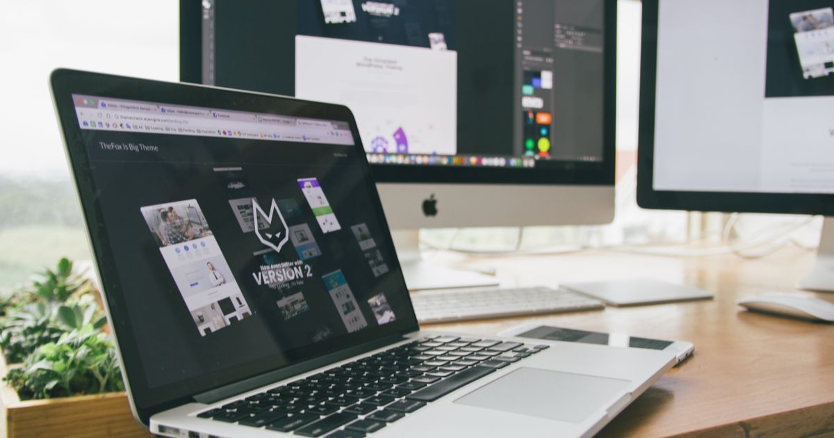 A MacBook Pro on a wooden desk displaying a website design mockup, with additional monitors showing design software in the background