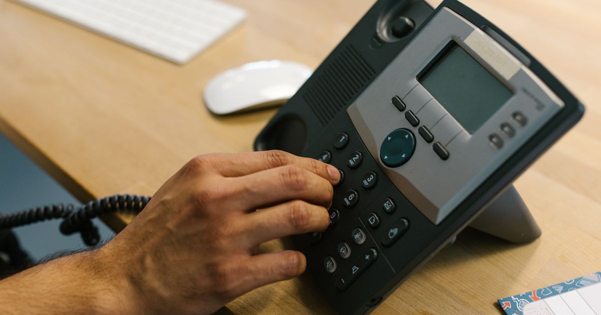 Close-up of a hand dialing an office desk phone next to a keyboard and mouse